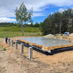 Marek Kruszczyński - Zalany betonem fundament z drewnianym szalunkiem i izolacją, widoczne słupki, w tle łąka i las, błękitne niebo z chmurami.