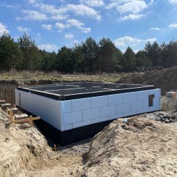 Marek Kruszczyński - Ocieplone fundamenty domu jednorodzinnego z widoczną izolacją przeciwwilgociową i termiczną, na tle wykopu i drzew. Etap budowy zrealizowany w słoneczny dzień.
