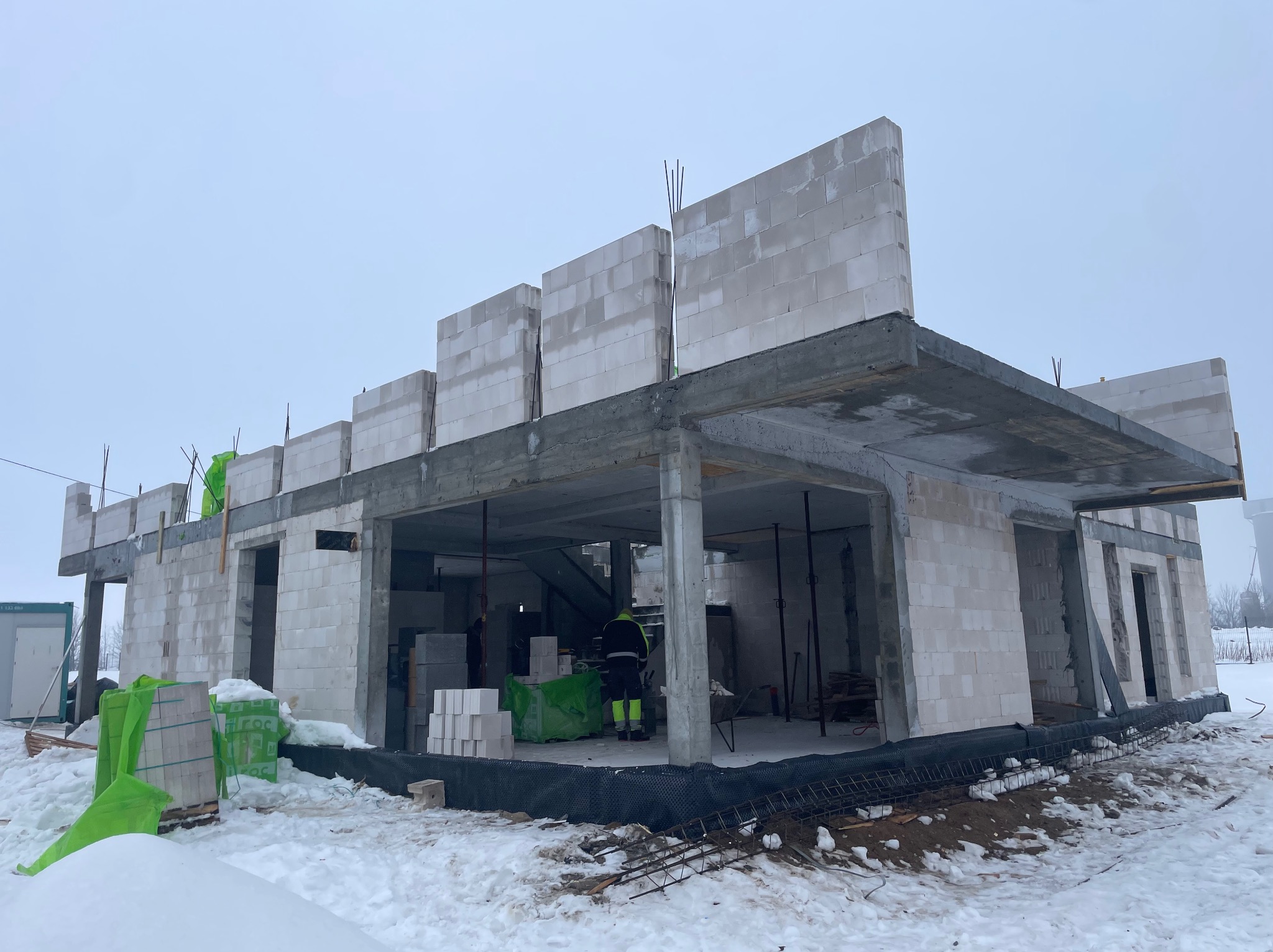 Niedokończony dom z betonu komórkowego w zimowej scenerii. Widoczne ściany parteru, betonowy strop i materiały budowlane na śniegu. Prace murarskie w toku.