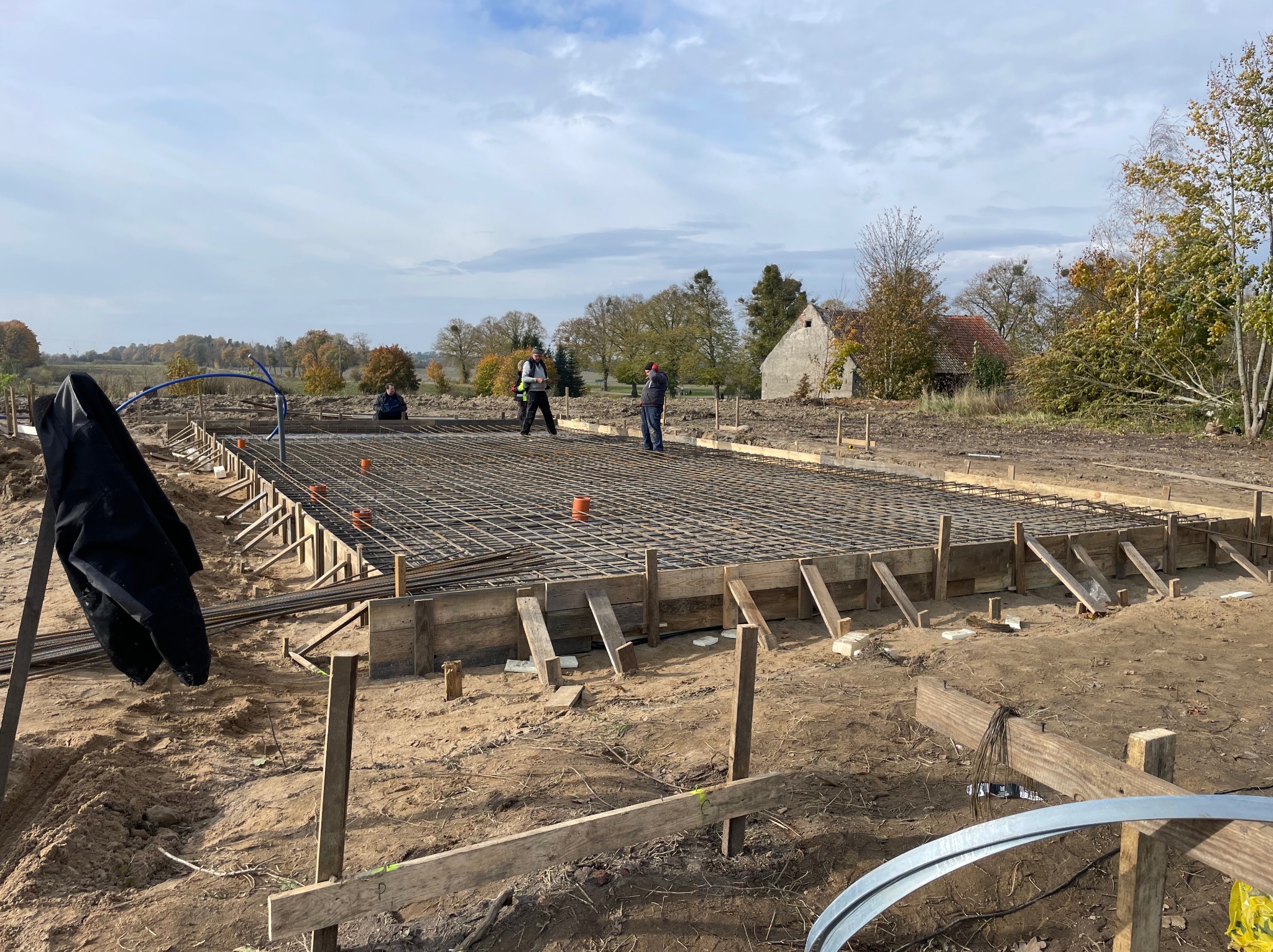 Zbrojenie fundamentów pod budowę domu jednorodzinnego. Widoczne drewniane szalunki, stalowe pręty zbrojeniowe i pracownicy na placu budowy. W tle zabudowania wiejskie.