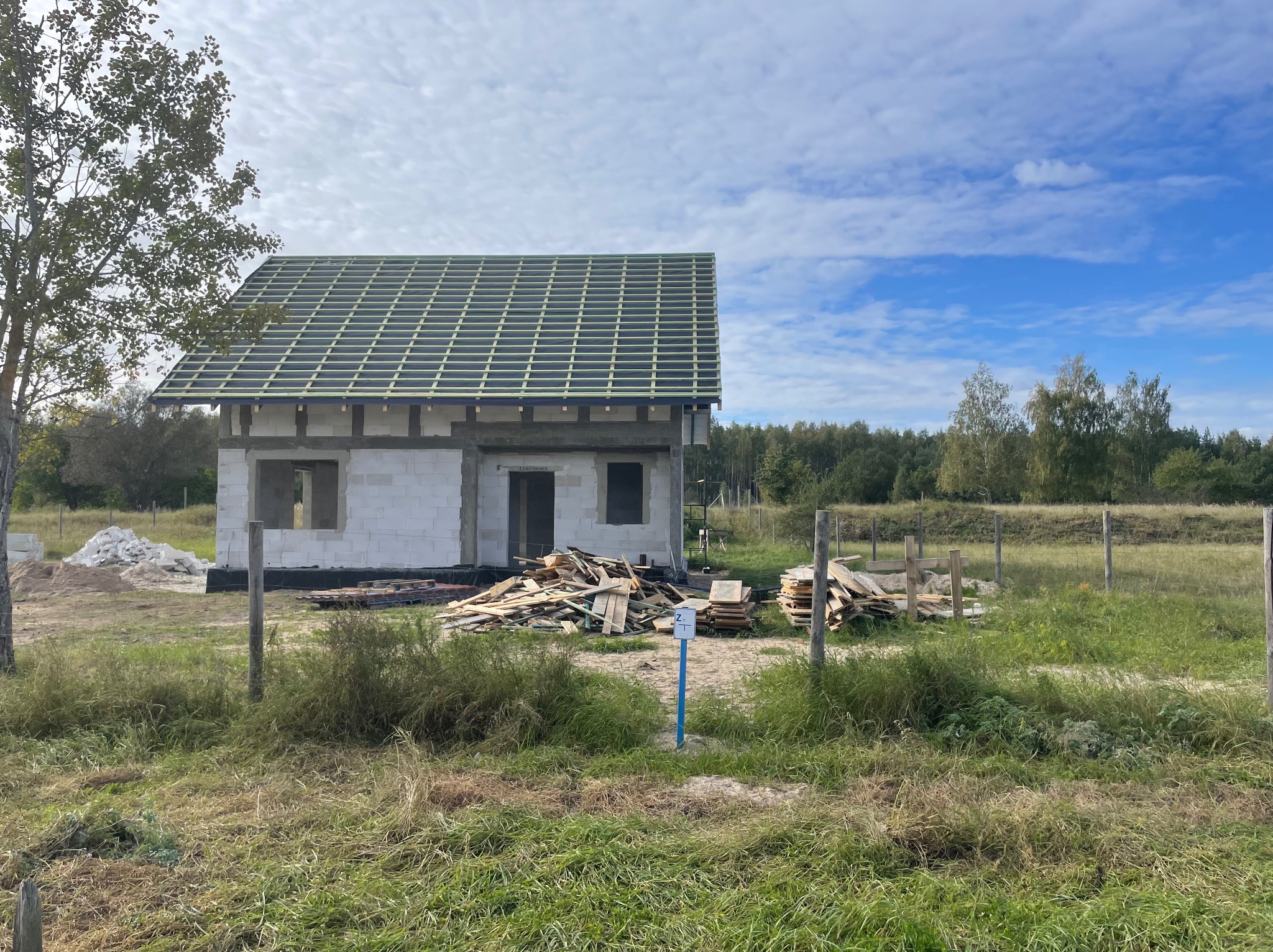 Niedokończony dom z białego pustaka, z zieloną konstrukcją dachu, otoczony trawą i drzewami. Na pierwszym planie stos desek i niebieski słupek.