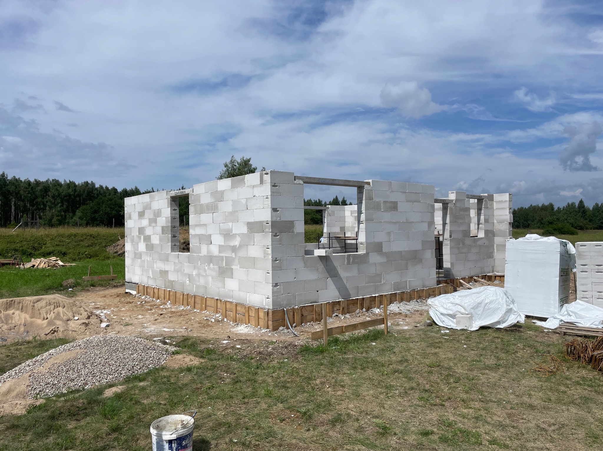 Wznoszenie ścian parterowego domu z białych bloczków, widoczne otwory okienne i drzwiowe, drewniane szalunki fundamentów, materiały budowlane na działce, błękitne niebo.