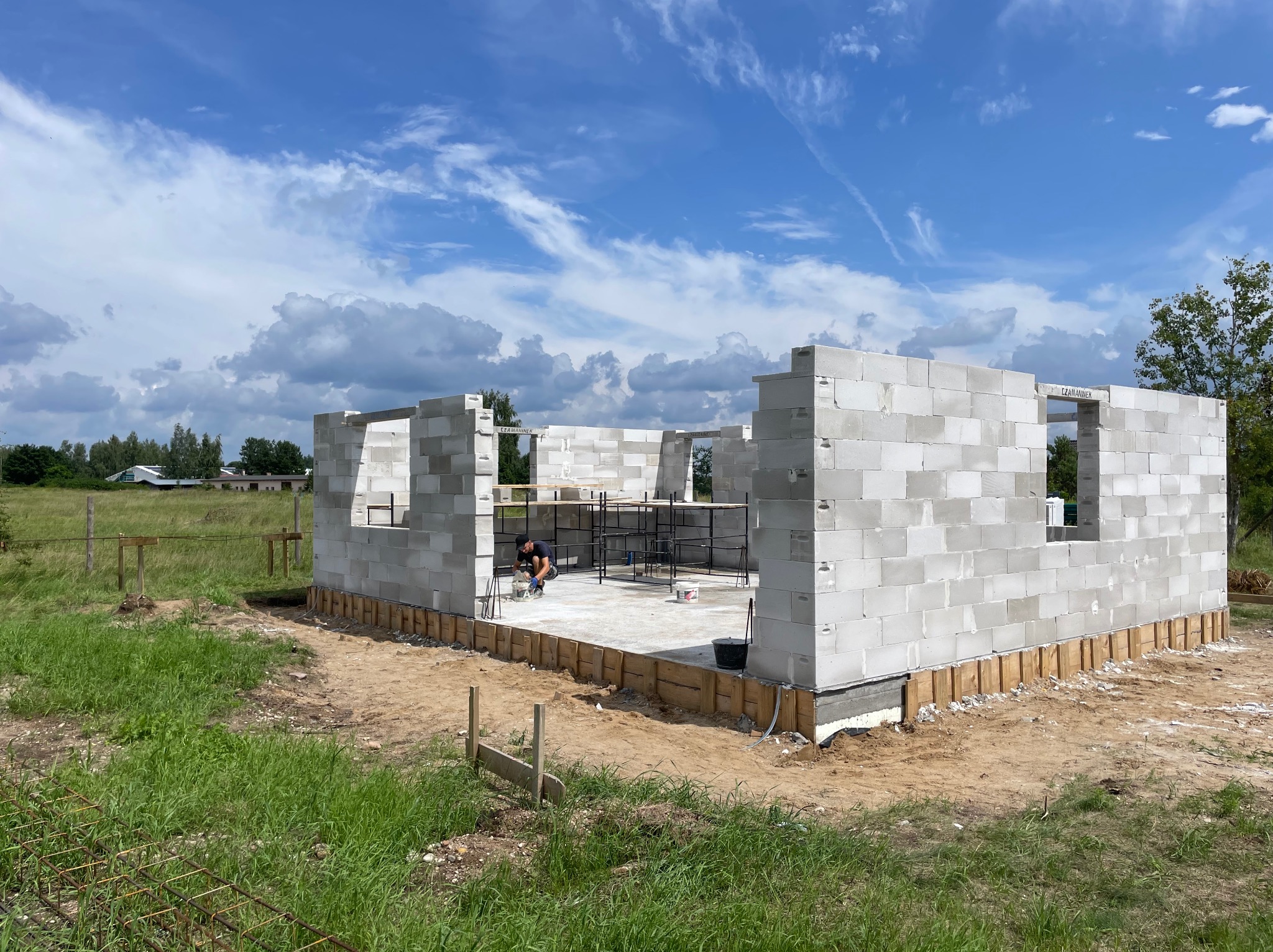 Budowa domu z białych bloczków: wzniesione ściany parteru, widoczny pracownik wewnątrz, drewniane szalunki fundamentów, błękitne niebo z chmurami w tle.
