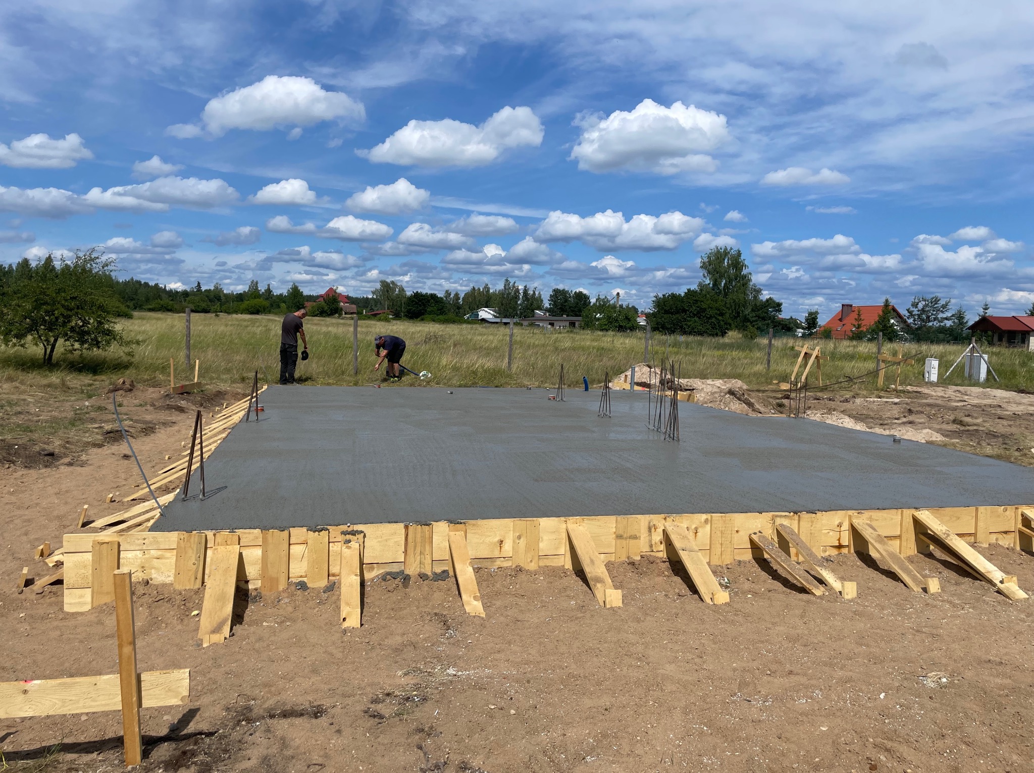 Zalany fundament pod budowę domu, drewniane szalunki, wystające pręty zbrojeniowe, dwóch pracowników w tle, błękitne niebo z chmurami.