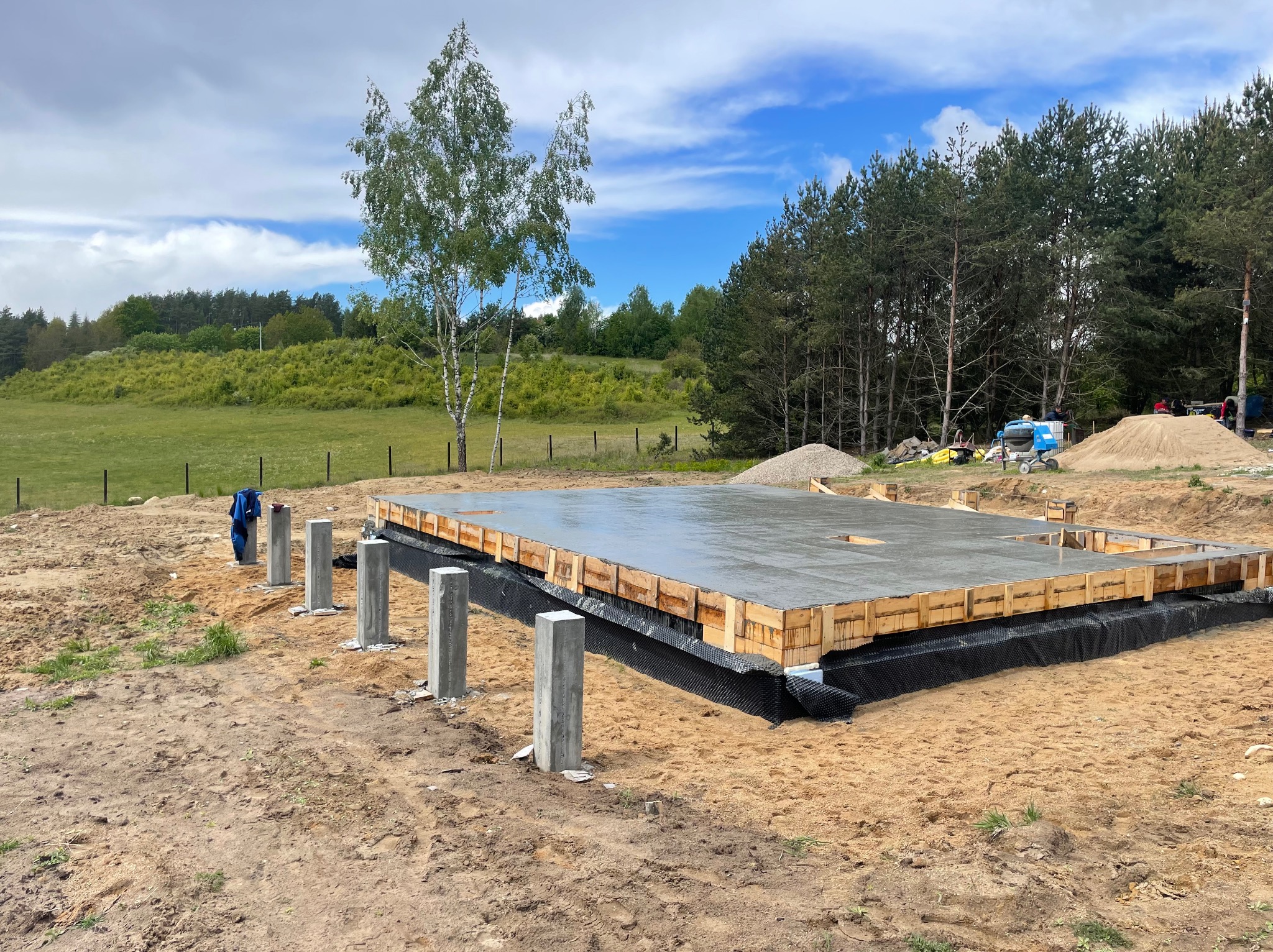 Zalany betonem fundament z drewnianym szalunkiem i izolacją, widoczne słupki, w tle łąka i las, błękitne niebo z chmurami.