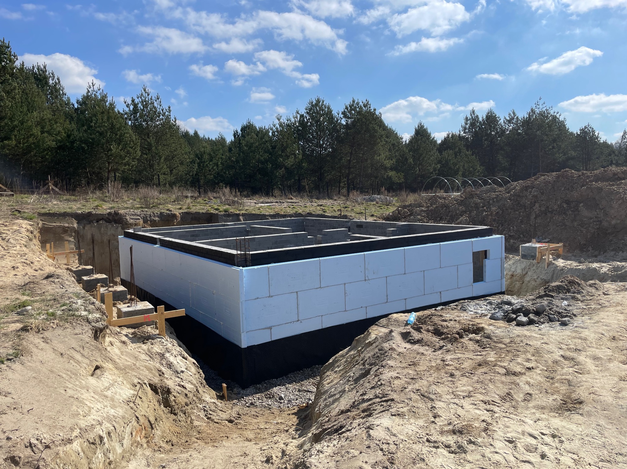 Ocieplone fundamenty domu jednorodzinnego z widoczną izolacją przeciwwilgociową i termiczną, na tle wykopu i drzew. Etap budowy zrealizowany w słoneczny dzień.