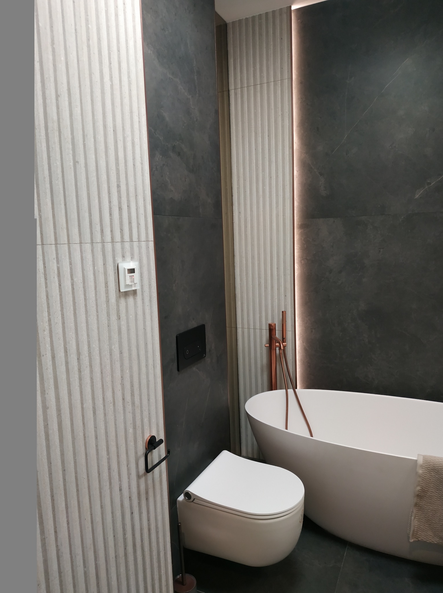 Nowoczesna łazienka z wolnostojącą wanną, szarą płytką imitującą kamień i dekoracyjną, pionową płytką w paski. Armatura w kolorze miedzi i podświetlenie LED dodają elegancji.