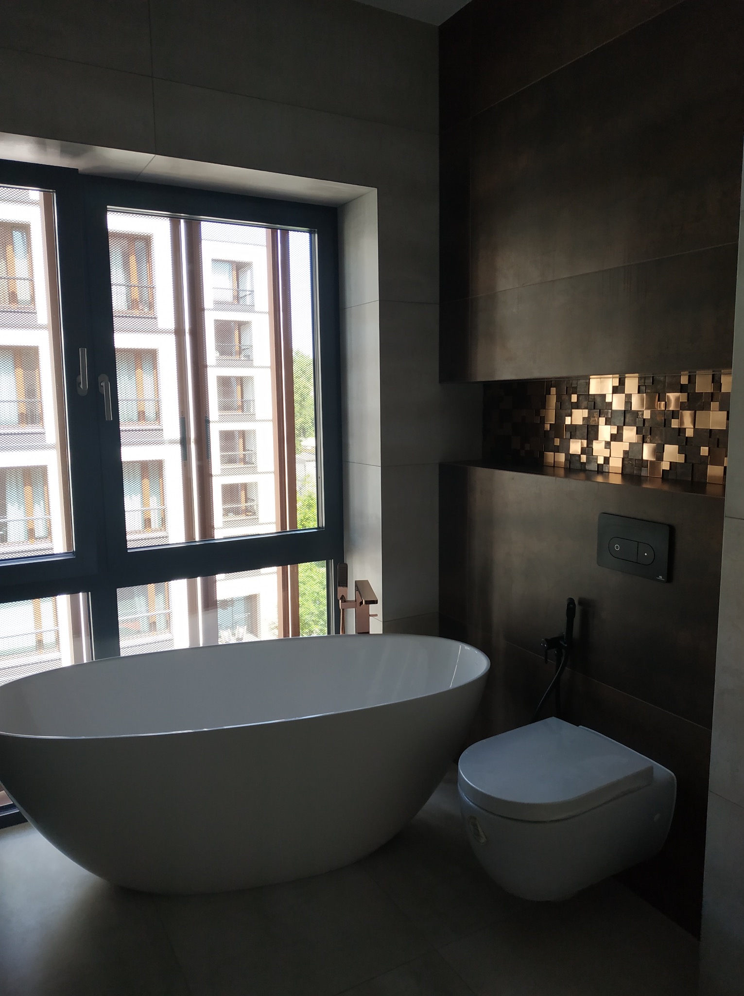 Nowoczesna łazienka z wolnostojącą wanną przy oknie i podwieszaną toaletą na tle ściany z dekoracyjną wnęką z mozaiką. Minimalistyczny design.