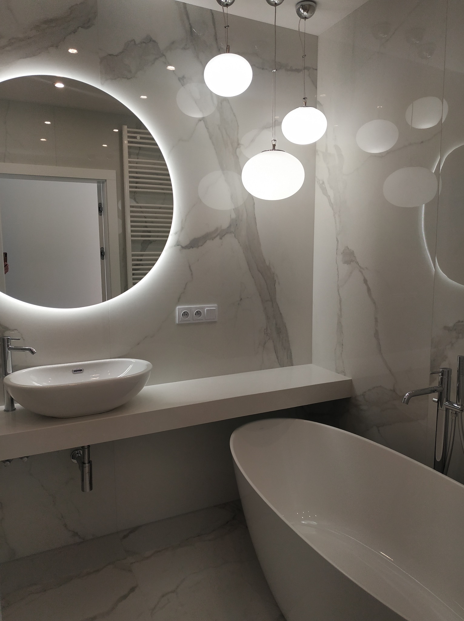 Nowoczesna łazienka z okrągłym lustrem LED, marmurowymi płytkami i wolnostojącą wanną. Minimalistyczny design, subtelne oświetlenie, armatura w kolorze chromu.