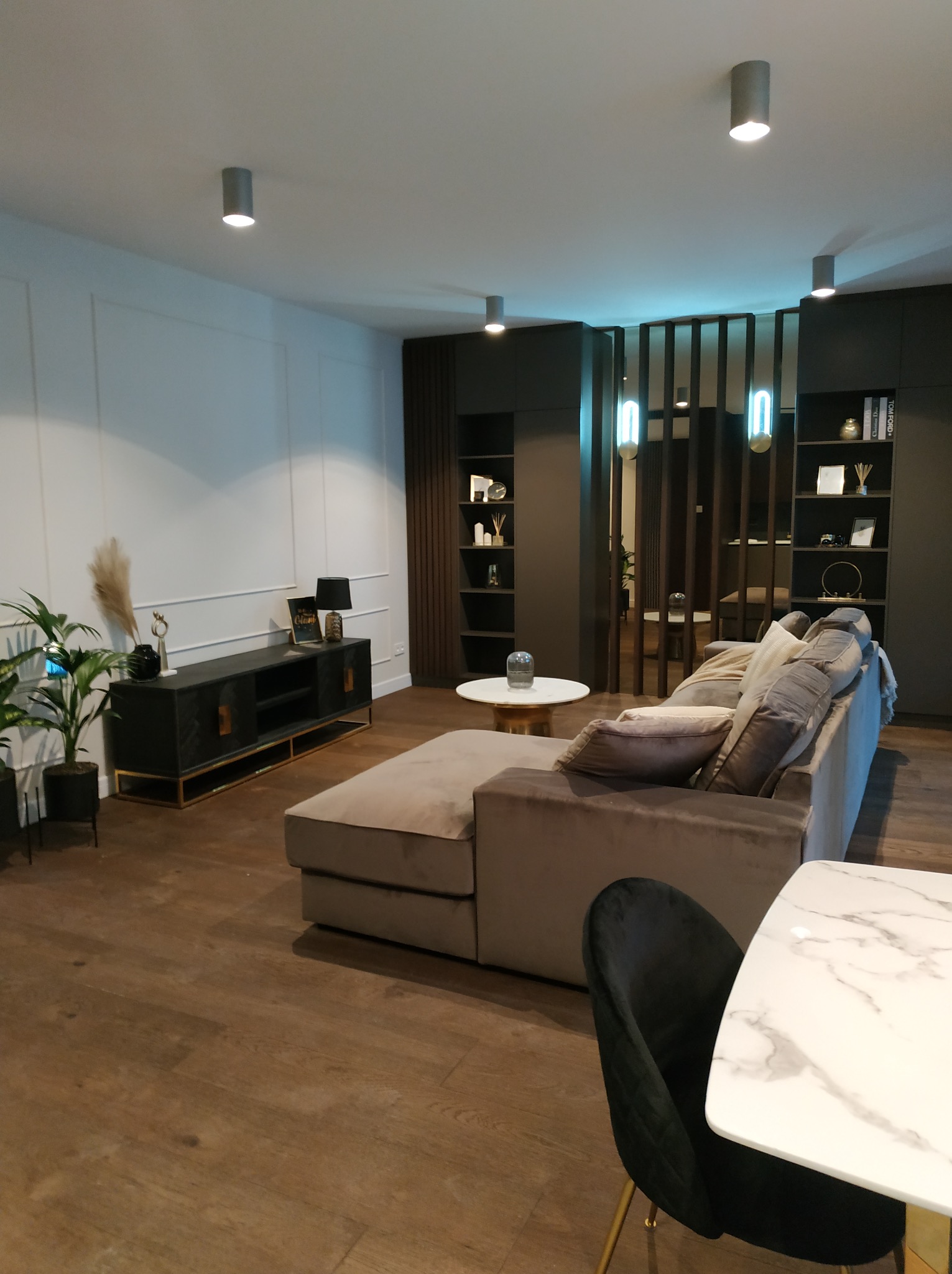 Nowoczesny salon z beżową sofą, drewnianą podłogą i czarnymi meblami. Białe ściany z dekoracyjnymi listwami. Minimalistyczne oświetlenie sufitowe.