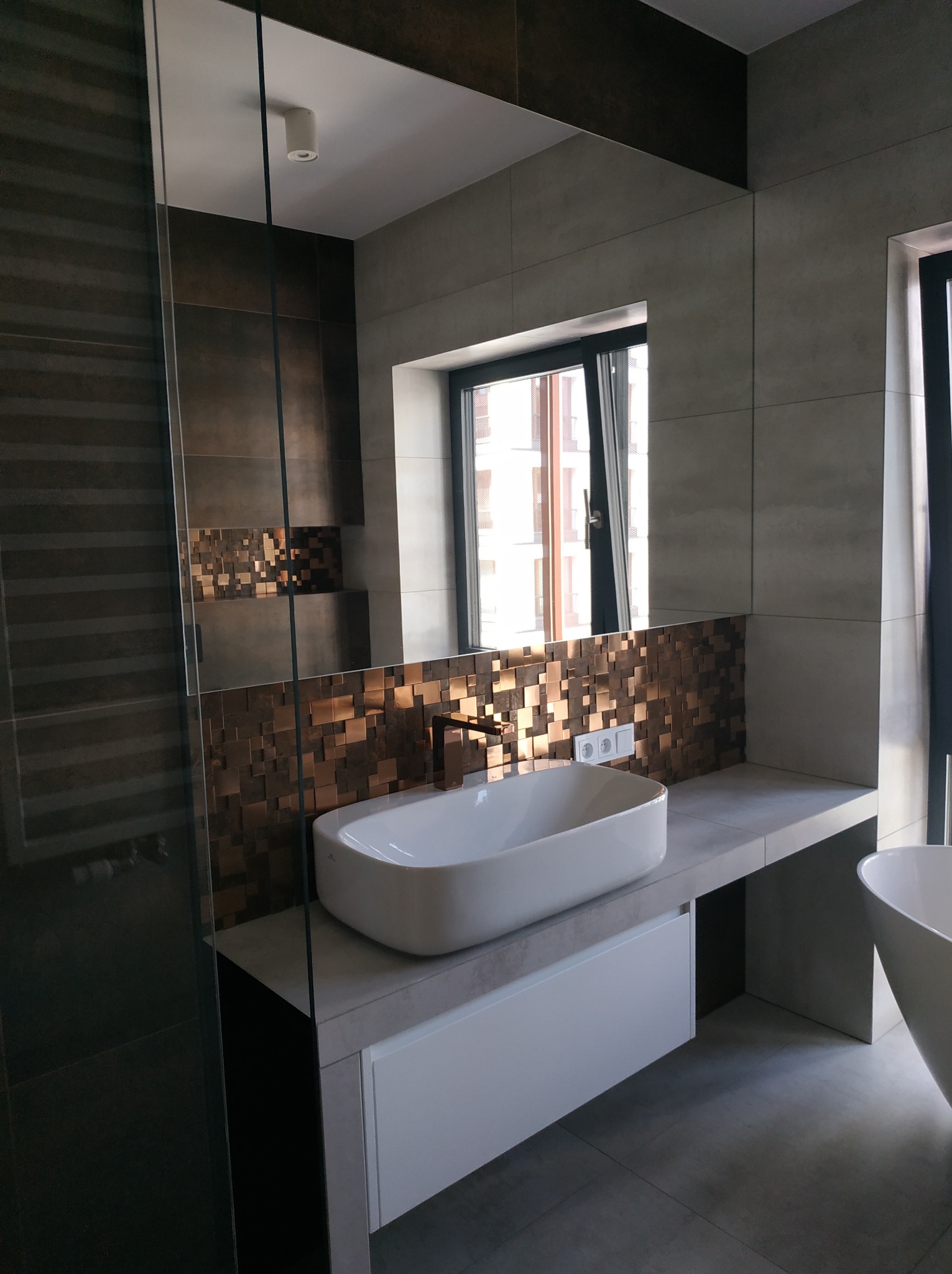 Nowoczesna łazienka z umywalką nablatową, mozaiką z metalicznych kostek i dużym lustrem. Minimalistyczny design, stonowana kolorystyka, widok na okno.