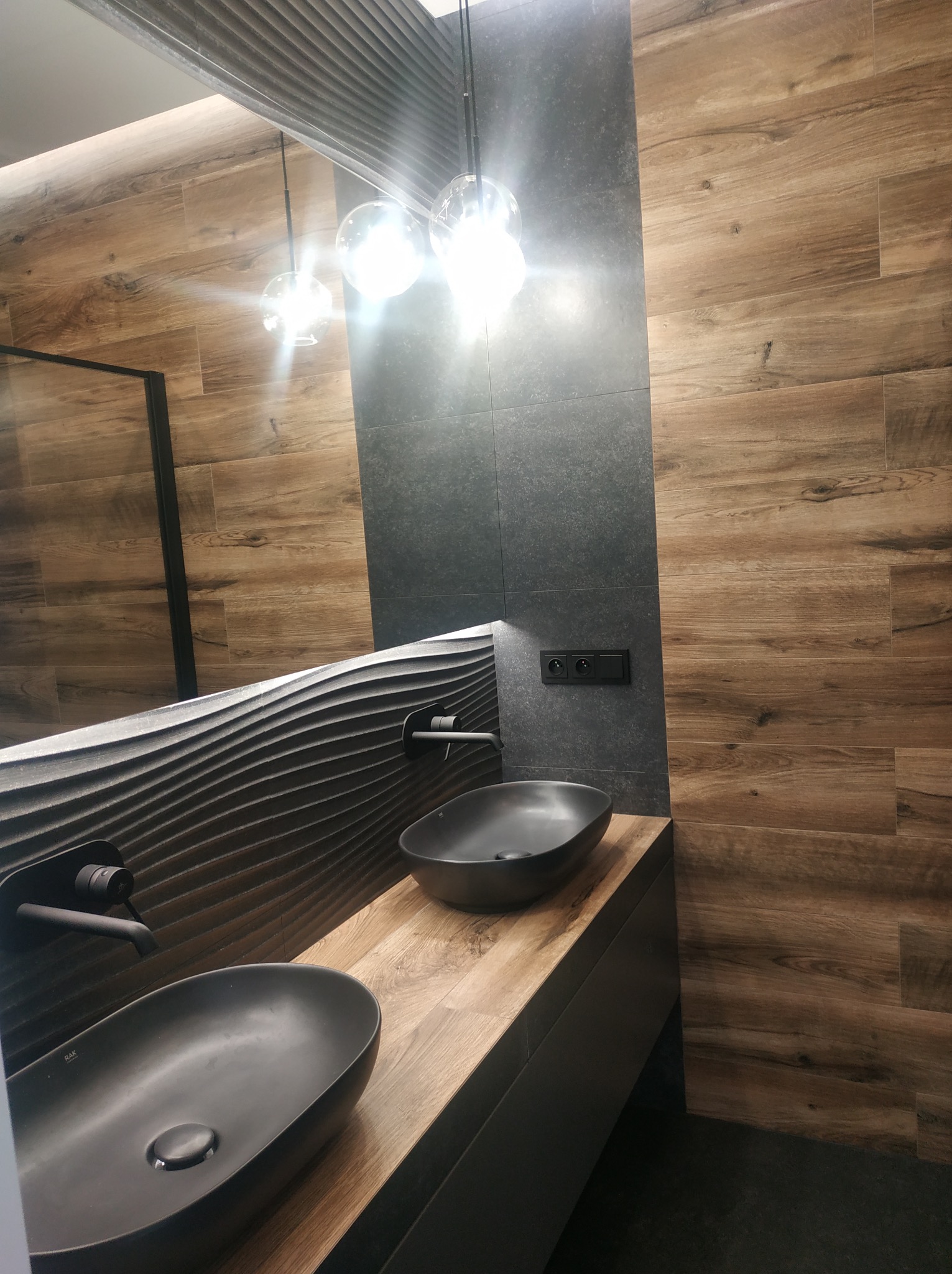 Nowoczesna łazienka z drewnopodobnymi płytkami, czarną armaturą i umywalkami nablatowymi. Dekoracyjna ściana z efektem falowania. Minimalistyczne oświetlenie.