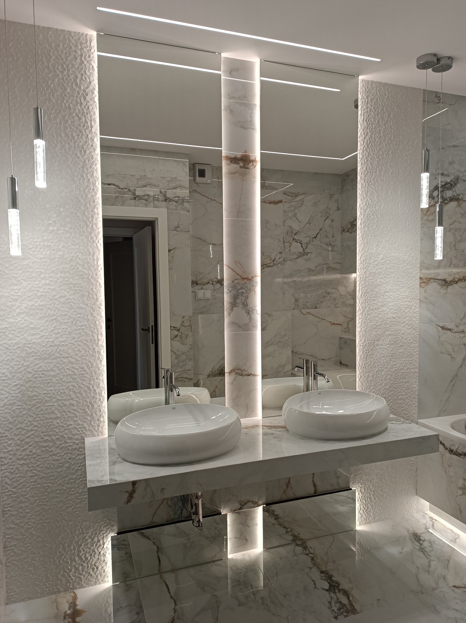 Nowoczesna łazienka z marmurowymi płytkami, podwieszanymi umywalkami i oświetleniem LED. Lustro z wbudowanym światłem. Teksturowana ściana dodaje unikalnego charakteru.