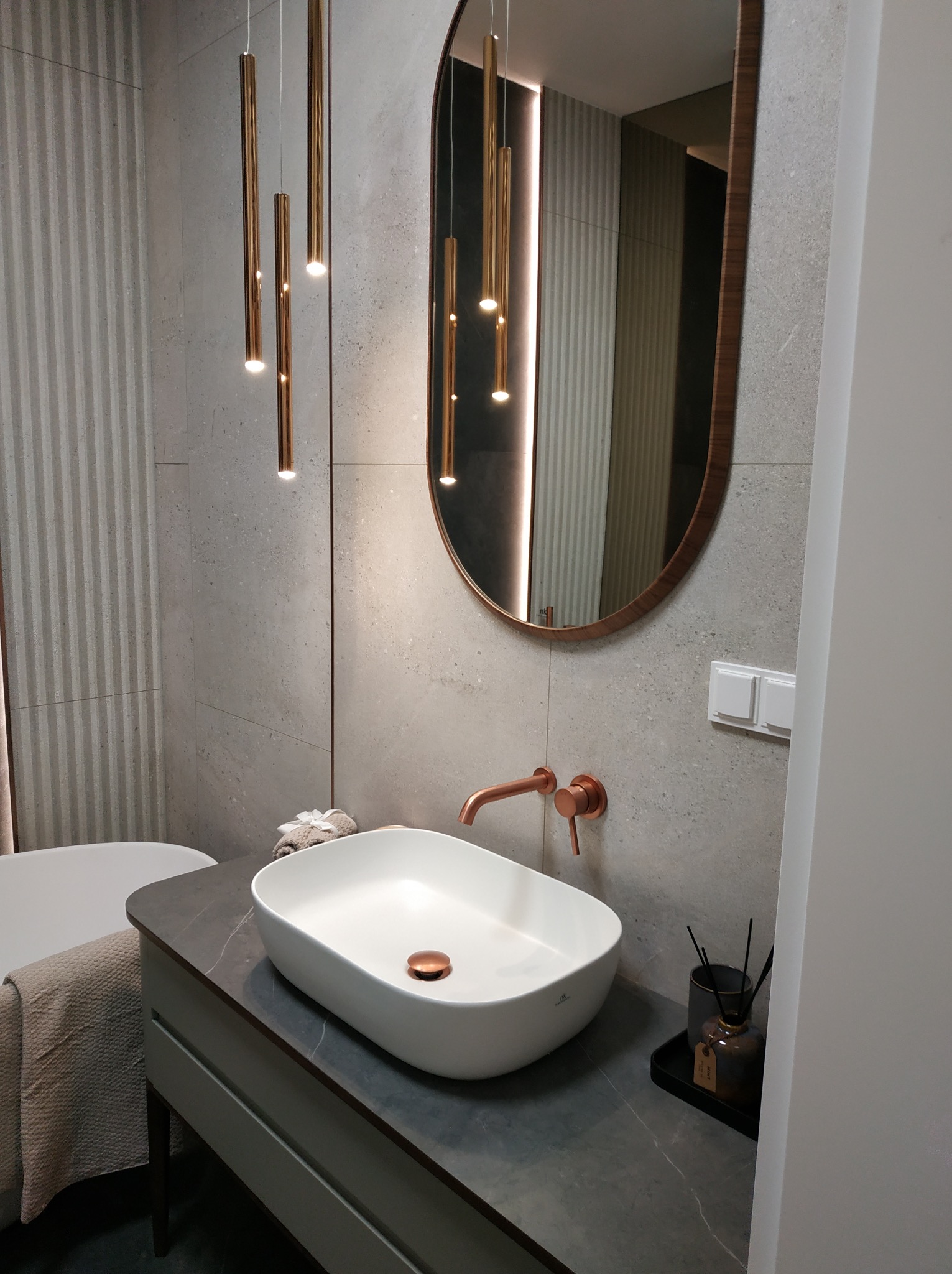 Nowoczesna łazienka z owalną umywalką nablatową, baterią w kolorze miedzi i podłużnym lustrem. Na ścianie szare płytki z delikatnym wzorem. Wiszące lampy tuby.