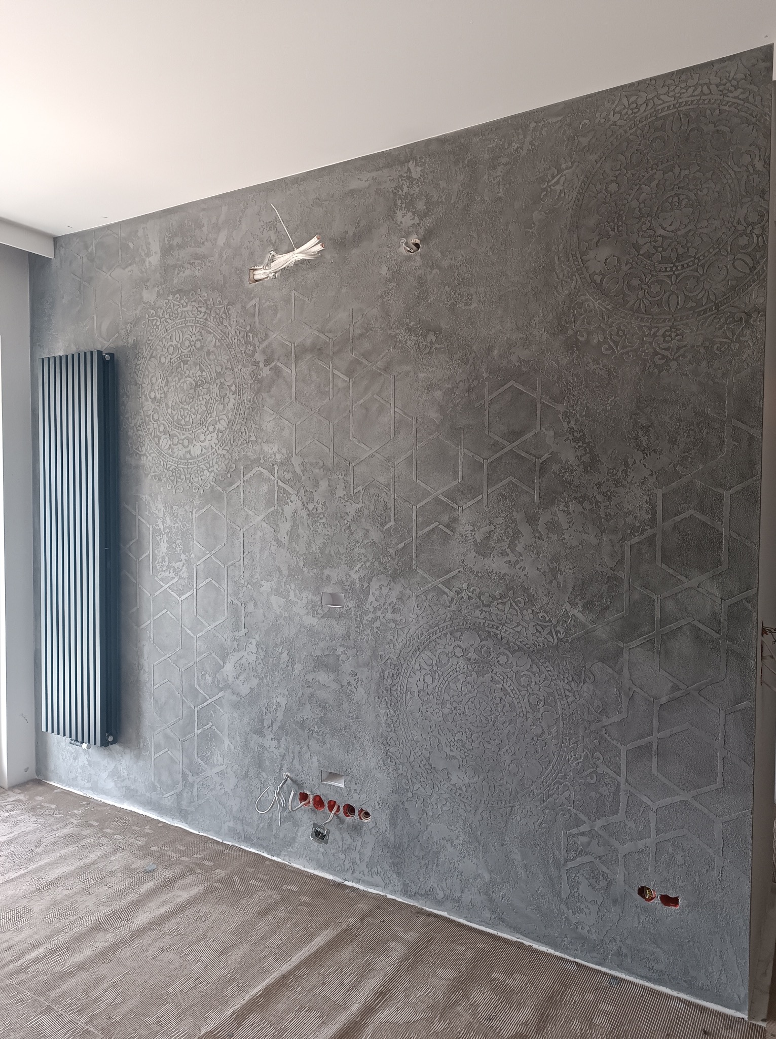 Dekoracyjna ściana w odcieniach szarości z geometrycznymi i okrągłymi wzorami. Widoczne gniazdka elektryczne i grzejnik. Podłoga zabezpieczona folią malarską.
