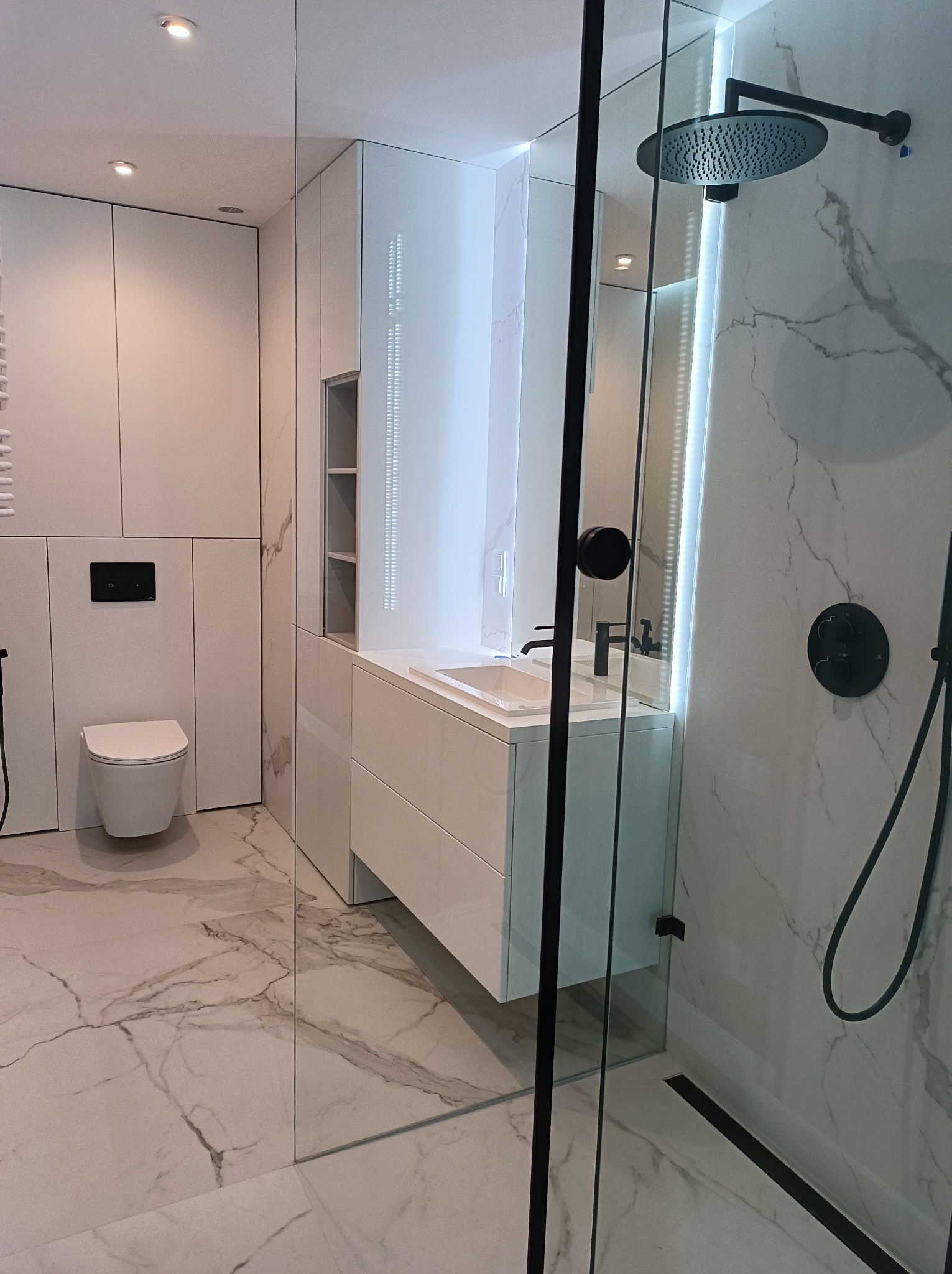 Nowoczesna łazienka z kabiną typu walk-in, białą armaturą i marmurową posadzką. Minimalistyczny design z czarnymi akcentami i oświetleniem LED.