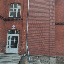 Oleksandr Soboda - Ceglana elewacja budynku z nową białą stolarką okienną i drzwiową. Widoczne schody wejściowe i rynna. Detal architektoniczny po renowacji.