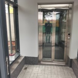 Oleksandr Soboda - Nowoczesna winda osobowa z metalową ramą i szklanymi drzwiami przesuwnymi, widoczna z zewnątrz budynku. Na podłodze szara kostka brukowa.