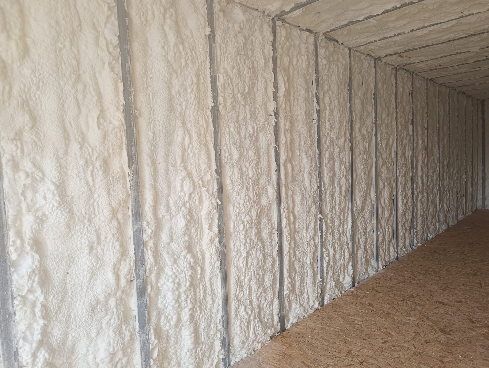 Ściana z metalowymi profilami wypełniona białą pianą izolacyjną, widok z dołu, ukazujący strukturę piany i drewnianą podłogę. Detal izolacji natryskowej.