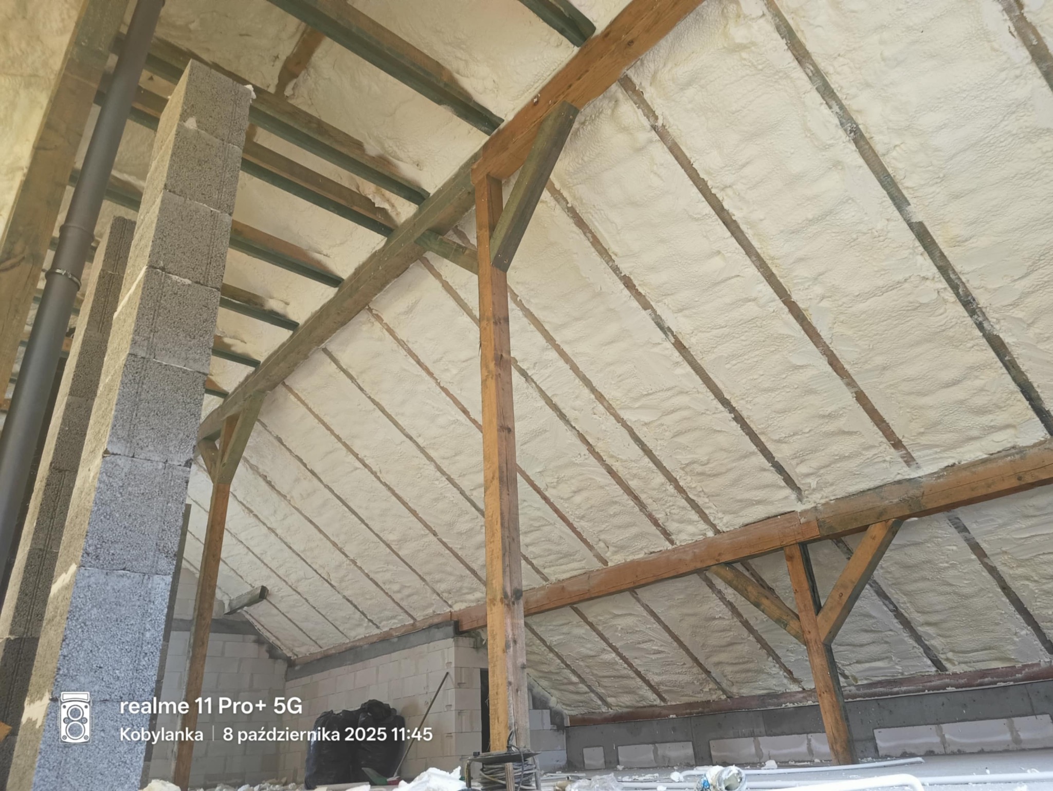 Poddasze z drewnianą konstrukcją dachu, pokryte grubą warstwą białej pianki izolacyjnej. Widoczne elementy konstrukcyjne i fragment ściany z bloczków.