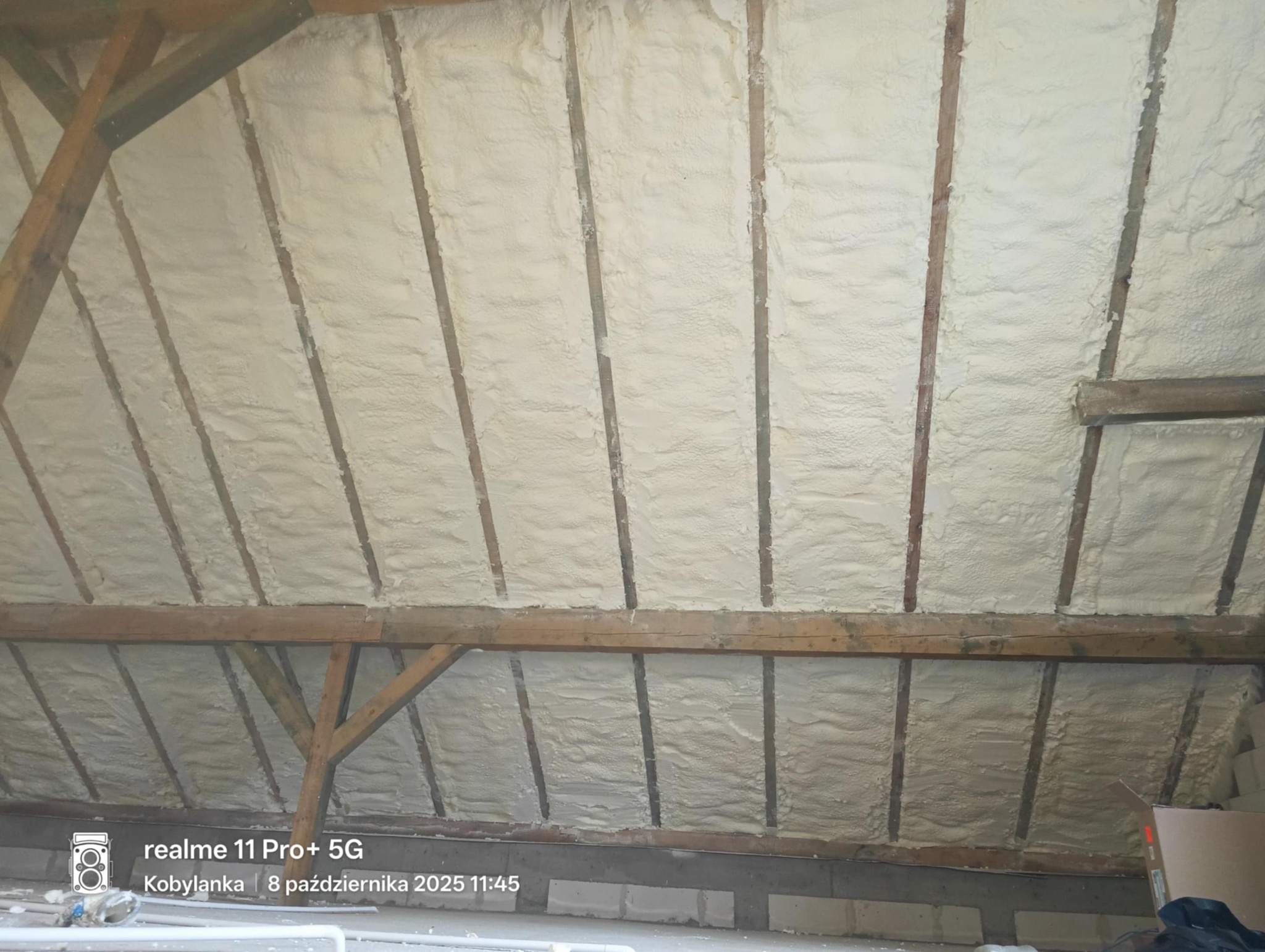 Poddasze z drewnianą konstrukcją pokryte warstwą jasnej pianki izolacyjnej, widoczne z dołu pod kątem. Elementy konstrukcyjne częściowo widoczne.
