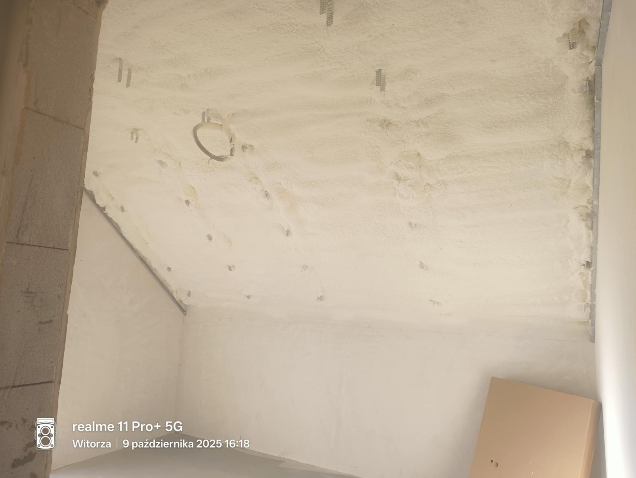 Poddasze po natrysku pianki izolacyjnej, widoczne elementy konstrukcyjne i otwór na oświetlenie. Biała, nierówna powierzchnia pianki pokrywa całą przestrzeń.