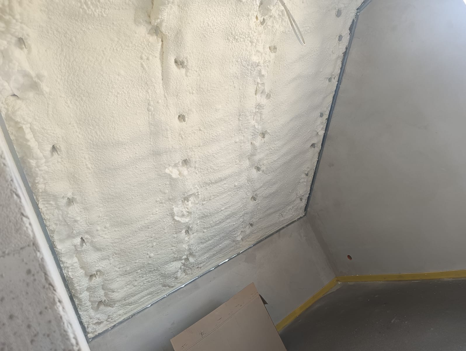 Poddasze po natrysku pianki PUR, widoczne punkty mocowania. Biała, porowata struktura izolacji wypełnia przestrzeń między krokwiami. Szara ściana i podłoga w tle.