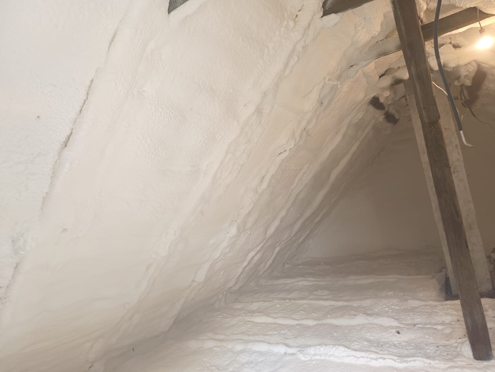 Poddasze z widoczną, grubą warstwą białej pianki izolacyjnej, pokrywającą skośne ściany i podłogę, z elementami drewnianej konstrukcji dachu.