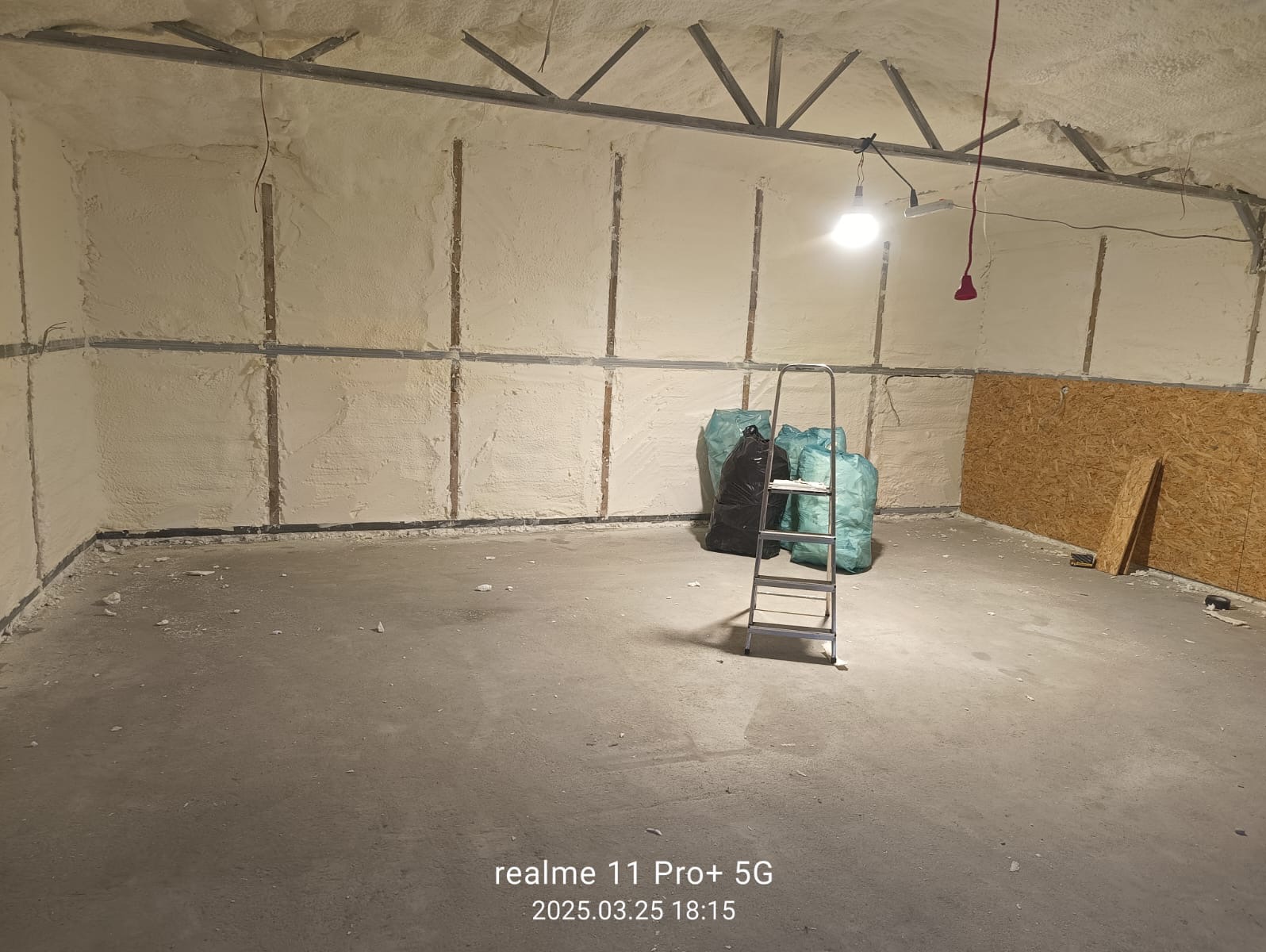 Poddasze po natrysku pianki PUR, widoczne worki z odpadami i drabina. Ściany i sufit pokryte jasną pianą, odsłonięta konstrukcja dachu. Częściowo widoczna płyta OSB.