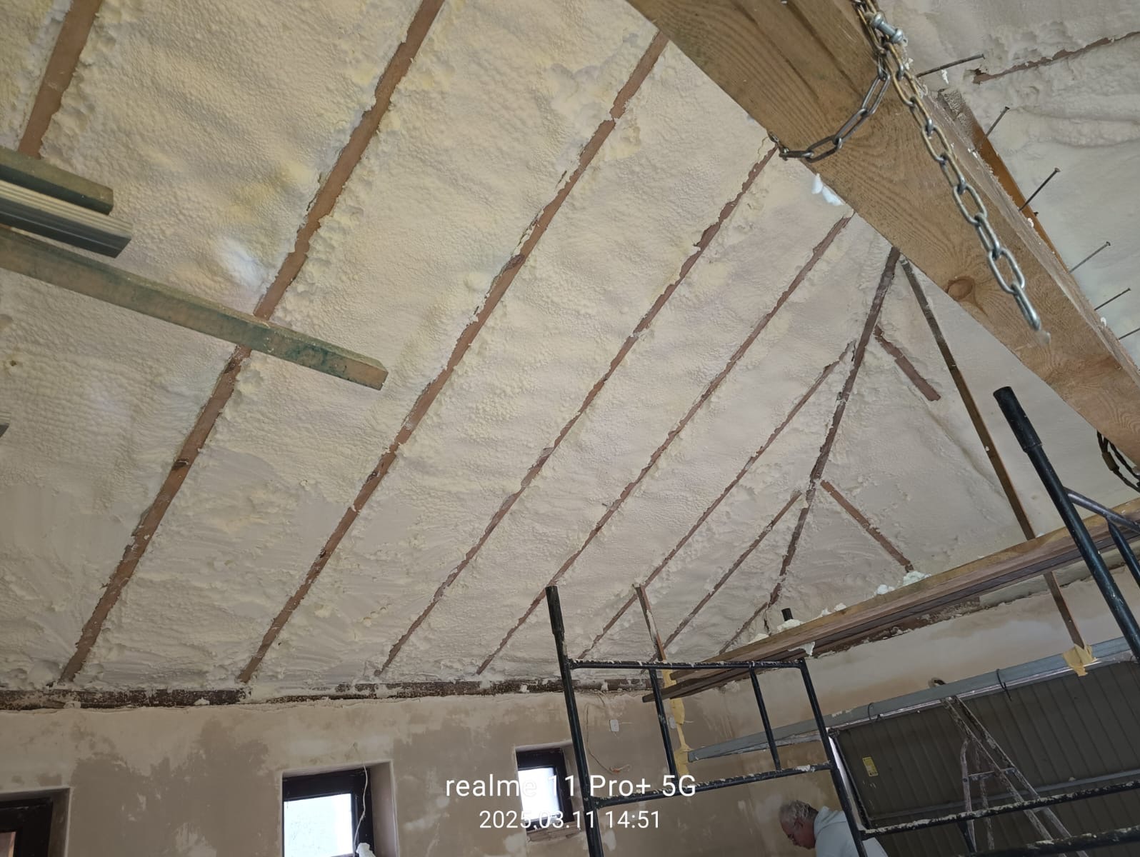 Poddasze z drewnianą konstrukcją pokryte warstwą jasnej pianki izolacyjnej, widoczne elementy rusztowania i fragment ściany z oknami.