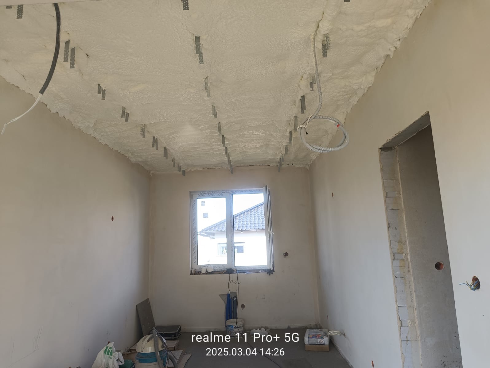 Poddasze po natrysku pianki, widoczne metalowe elementy montażowe. Ściany w surowym stanie, okno z widokiem na dach sąsiedniego budynku. Instalacja elektryczna na suficie.