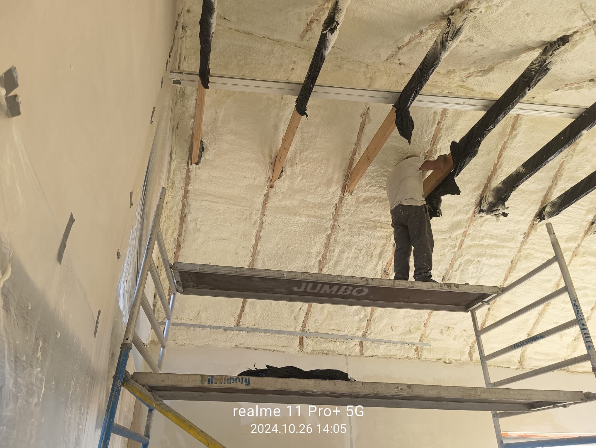 Pracownik na rusztowaniu aplikuje piankę PUR na poddaszu, widoczne drewniane elementy konstrukcyjne zabezpieczone folią. Detal: pianka o jednolitej strukturze.