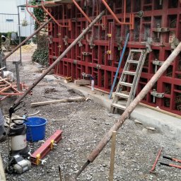 Kamil Chatya - Szczegółowy widok szalunku fundamentowego z czerwonymi panelami i metalowymi podporami na placu budowy, z narzędziami i drabiną w tle.