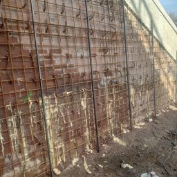 Kamil Chatya - Zbrojenie ściany z cegły z użyciem siatki stalowej i kotew. Widoczne ślady zaprawy i narzędzie budowlane na ziemi. Ujęcie z dołu, ukazujące etap prac budowlanych.