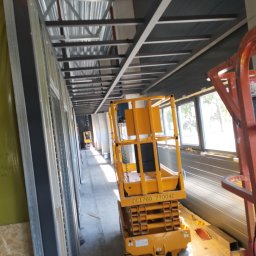 Kamil Chatya - Wnętrze budynku w trakcie budowy z widoczną konstrukcją stropu, ścianami z płyt i podnośnikiem koszowym. Widoczne okna i elementy konstrukcyjne.