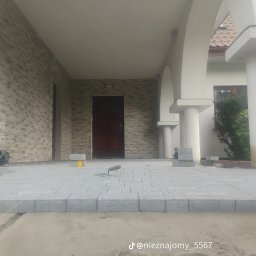 Bruk - Szara kostka brukowa ułożona na podjeździe domu z kolumnami. Widoczny fragment niezabrukowanej powierzchni i narzędzia pracy. Etap prac brukarskich.