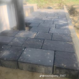 Bruk - Ciemnoszara kostka brukowa ułożona przy ścianie budynku, widoczne niedokończone fragmenty i rura spustowa. Krawężnik oddziela kostkę od ziemi.