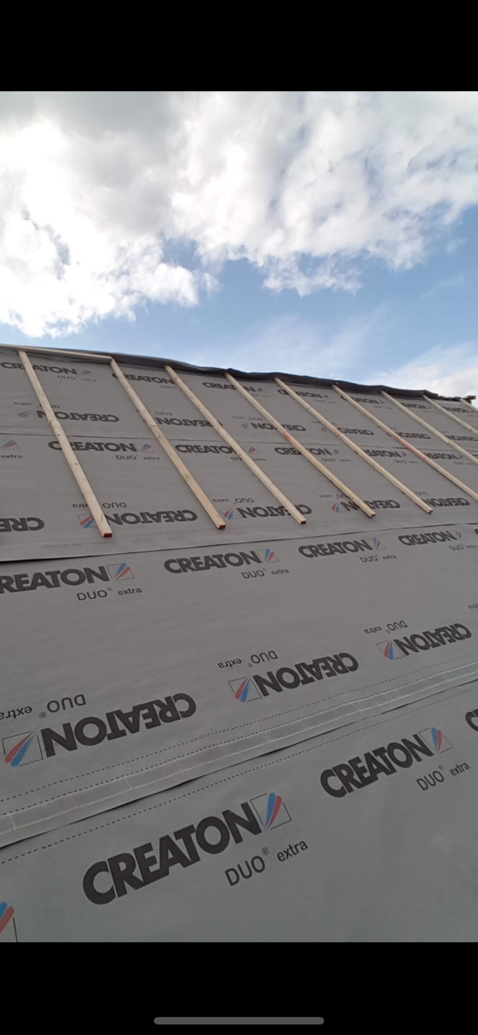 Dach w trakcie krycia: membrana CREATON Duo Extra i drewniane łaty pod dachówkę, widok z dołu na błękitne niebo z chmurami. Etap prac dekarskich.