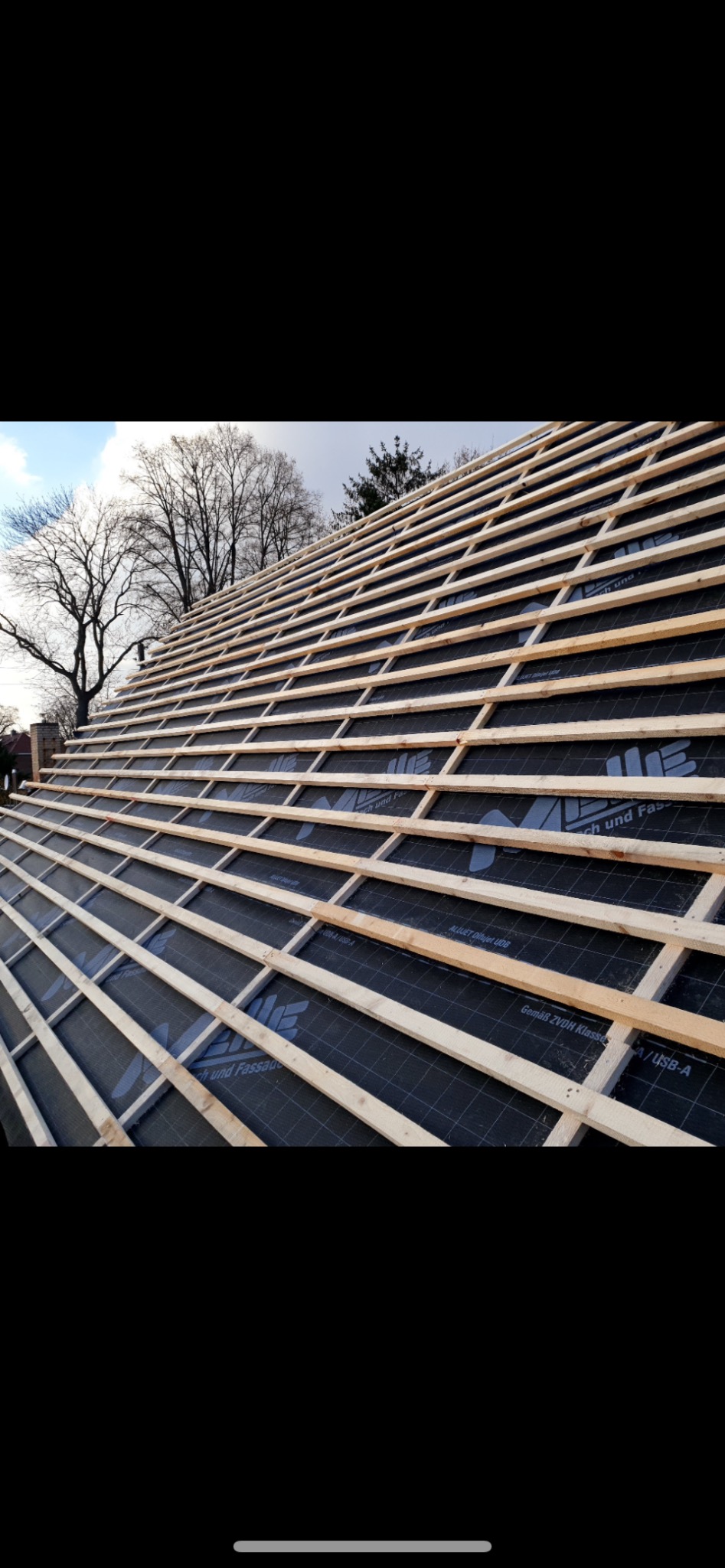 Konstrukcja dachu z drewnianymi łatami i membraną w tle drzewa i nieba. Widok z dołu na ukośną powierzchnię dachu w trakcie budowy.