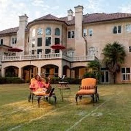 Młodszy Doradca Klienta Biznesowego - Elewacja dwupiętrowego domu z jasną fasadą, balkonem i czerwonymi parasolami. Na trawniku siedzą dwie osoby na krzesłach.