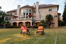 Elewacja dwupiętrowego domu z jasną fasadą, balkonem i czerwonymi parasolami. Na trawniku siedzą dwie osoby na krzesłach.