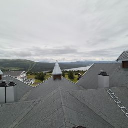 Ww Dachy - Szary dach z kominami i drabinką dekarską na tle krajobrazu z jeziorem i górami w pochmurny dzień. Widok z góry.