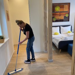 Mirosław Kaproń - Kobieta w dżinsach i czarnej koszulce myje podłogę mopem w pokoju z łóżkiem i obrazem. Widoczne drewniane kolumny i okno z rośliną.