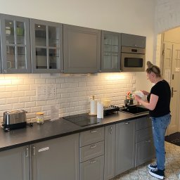 Mirosław Kaproń - Kobieta w kuchni w Sztum z szarymi szafkami i wzorzystą podłogą, myje naczynia przy zlewie. Widoczne akcesoria kuchenne i białe płytki na ścianie.