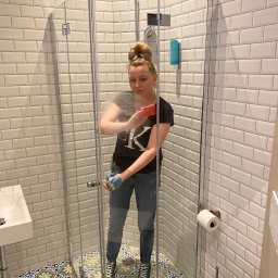 Mirosław Kaproń - Kobieta w jeansach i czarnej koszulce czyści kabinę prysznicową w łazience z białymi płytkami i wzorzystą podłogą, używając gąbki i płynu.