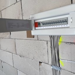 Kacper Kniter - Biała rozdzielnica elektryczna z 14 modułami i wyłącznikiem różnicowoprądowym, zamontowana na ścianie z bloczków, z widocznymi etykietami obwodów i ułożonymi kablami w bruździe.