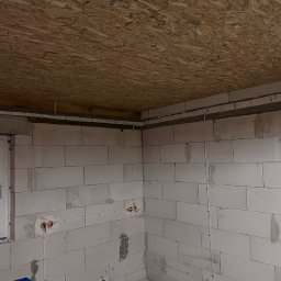 Kacper Kniter - Narożnik pomieszczenia w trakcie budowy z widoczną instalacją elektryczną: białe peszle na ścianach z bloczków i suficie z płyt OSB, z czerwonymi puszkami.