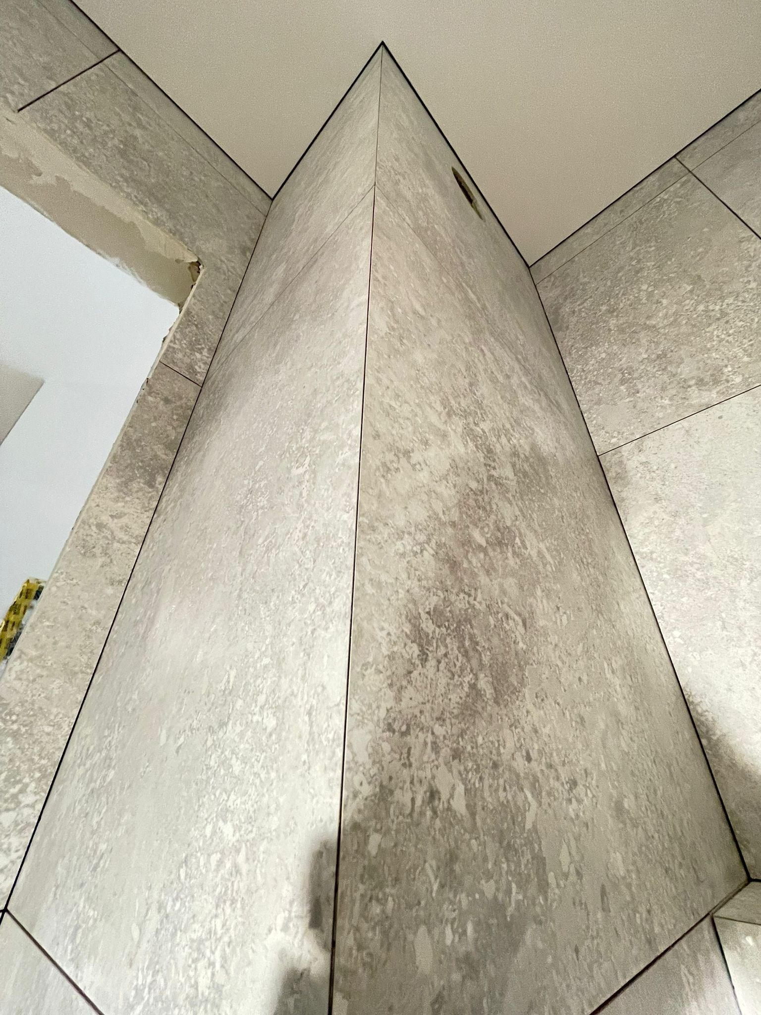 Ściana wyłożona szarymi, matowymi płytkami imitującymi kamień, z widocznymi fugami i otworem na instalację. Ujęcie z dołu podkreśla geometrię narożnika.