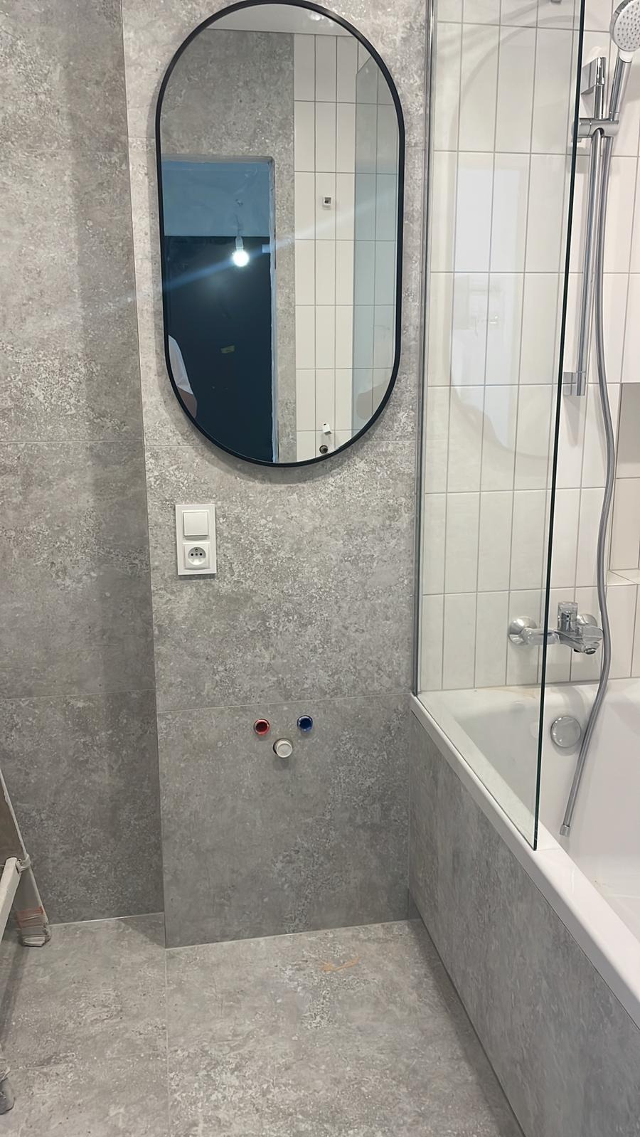 Nowoczesna łazienka po remoncie: szare płytki imitujące kamień, owalne lustro w czarnej ramie, biała wanna z szklanym parawanem i białe, wąskie płytki na ścianie.