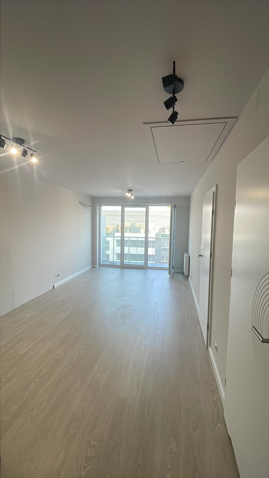 Jasne, przestronne wnętrze po remoncie z panelami podłogowymi, białymi ścianami i drzwiami balkonowymi. Widoczne oświetlenie sufitowe.